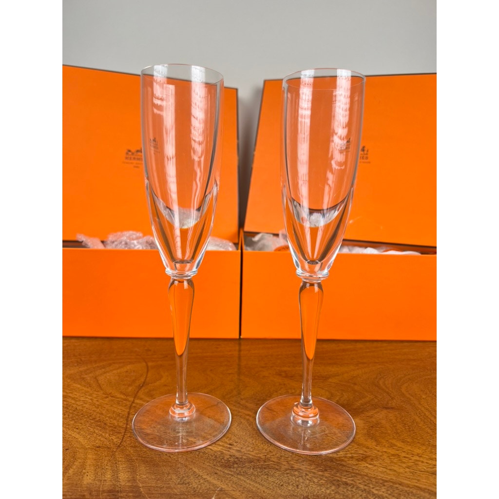 A set of eight Hermès Crystal Champagne Flutes by Saint Louis France (10.5") stands side by side on a wooden surface, their elegant glass surfaces reflecting light with Hermès’ signature orange boxes in the background.