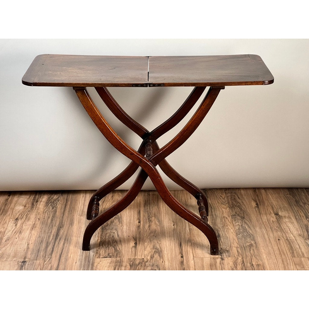 An Antique English Regency Mahogany Folding Coaching or Sporting Table (c. 1825–1845) with a rectangular top and curved X-shaped legs is set on a wood floor against a plain, light-colored wall.