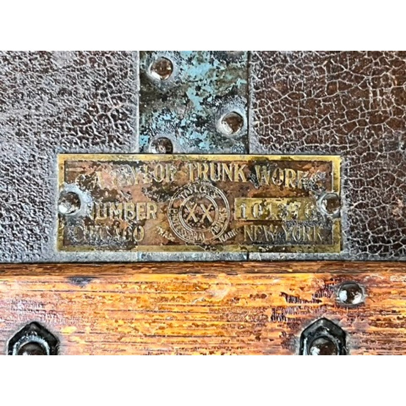 A close-up of a worn brass nameplate on a CA Taylor Touring Trunk owned by John B. Cordaro, Paul Whiteman Orchestra reed player, c. 1930s, reading “Taylor Trunk Works, Chicago, New York,” riveted to aged leather and wood.