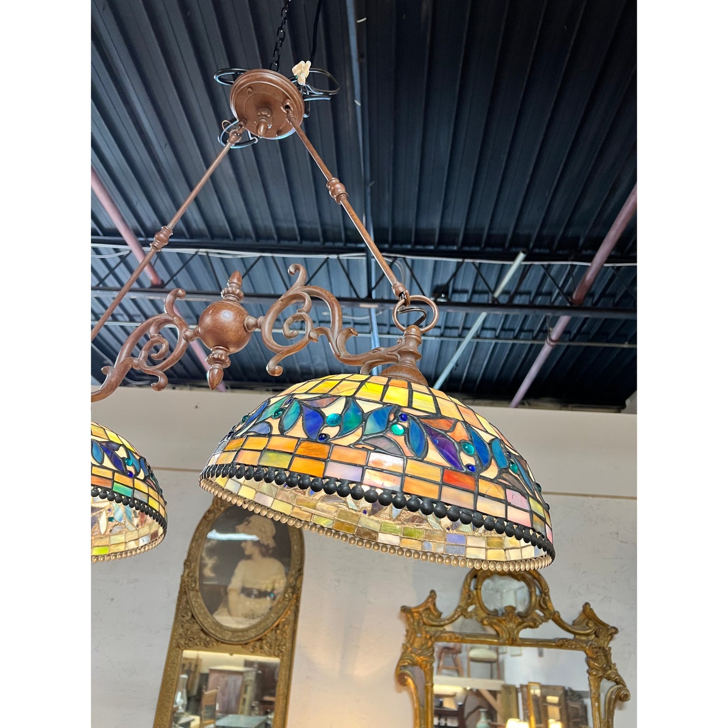 A Vintage Tiffany Style Leaded Stained Glass Double Pendant Billiard Island Light, featuring a colorful floral pattern, hangs from an ornate bronze fixture and is reflected in a mirror below on a white wall. Exposed black ceiling pipes are visible above. The brand is unknown.
