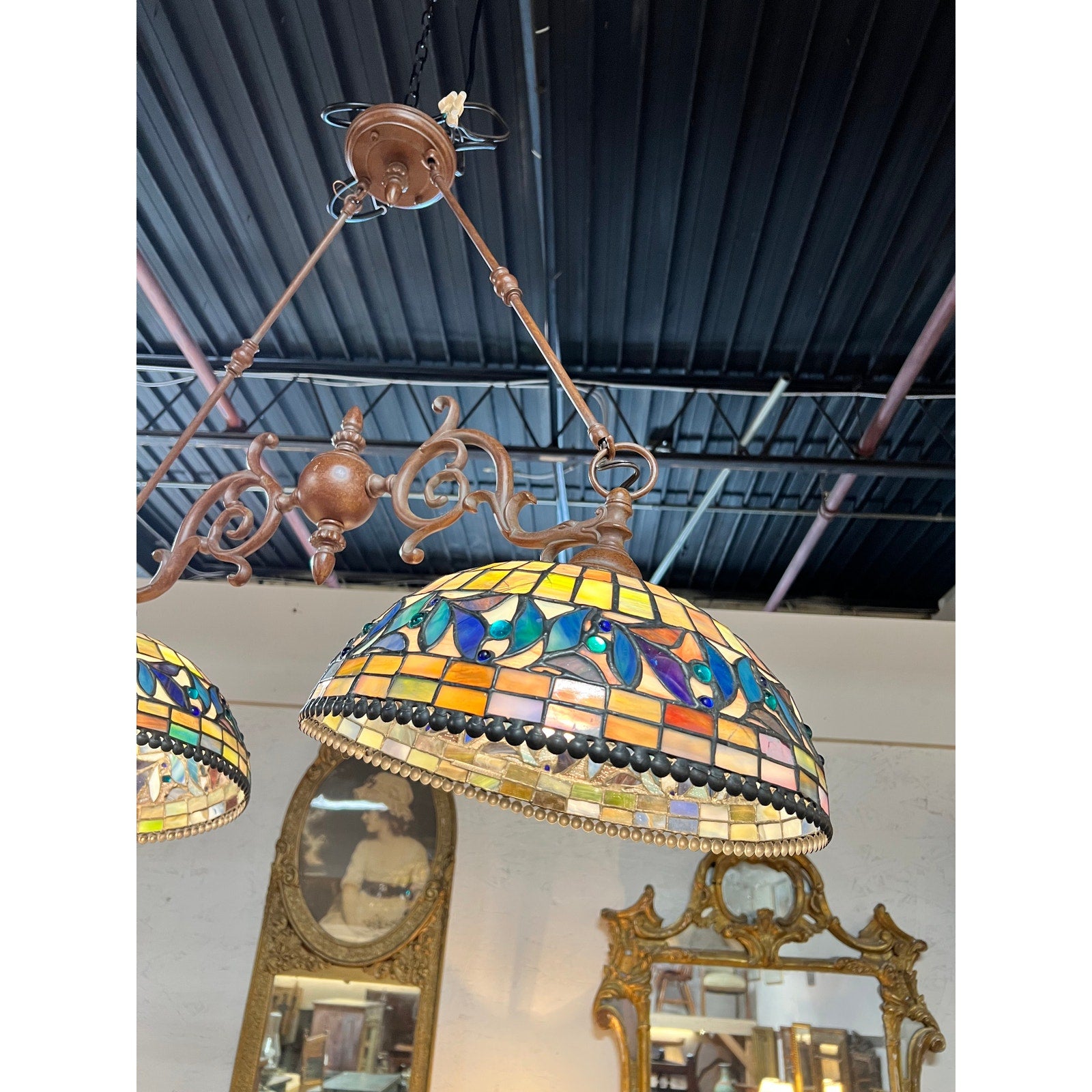 A Vintage Tiffany Style Leaded Stained Glass Double Pendant Billiard Island Light, featuring a colorful floral pattern, hangs from an ornate bronze fixture and is reflected in a mirror below on a white wall. Exposed black ceiling pipes are visible above. The brand is unknown.