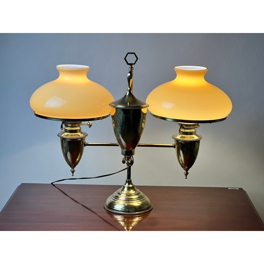 The Antique Bradley and Hubbard Brass Double-arm Student Lamp with Carmel Shades features warm glowing shades, classic double arms, and polished brass, echoing timeless Bradley and Hubbard craftsmanship on a wooden surface.