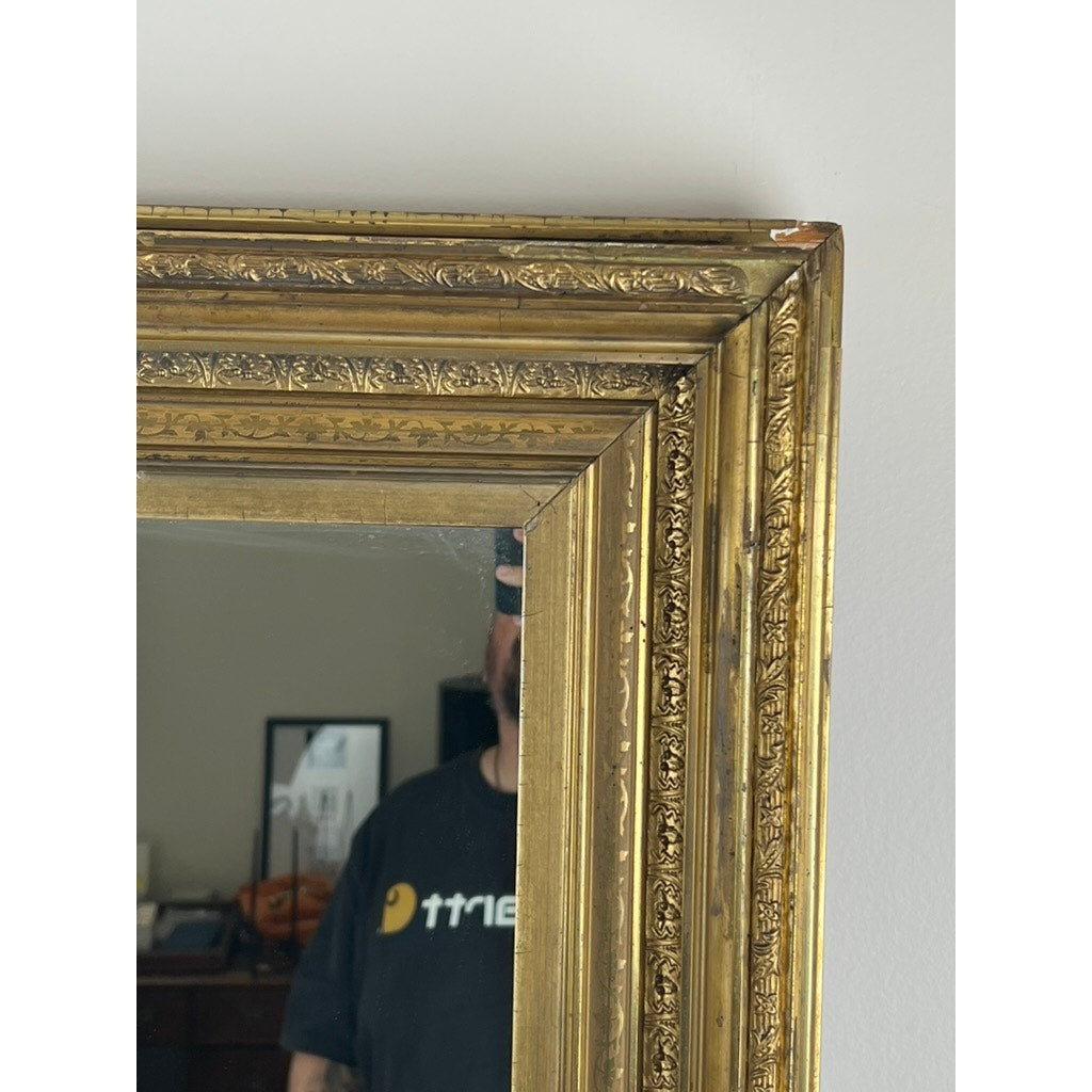 Close-up of the top left corner of a Fine Antique 19th Century Gilt Wood Mirror 39 x 34. A person wearing a black shirt with white text is reflected, with part of the wall and a picture visible in the background. Brand: None/Unknown.