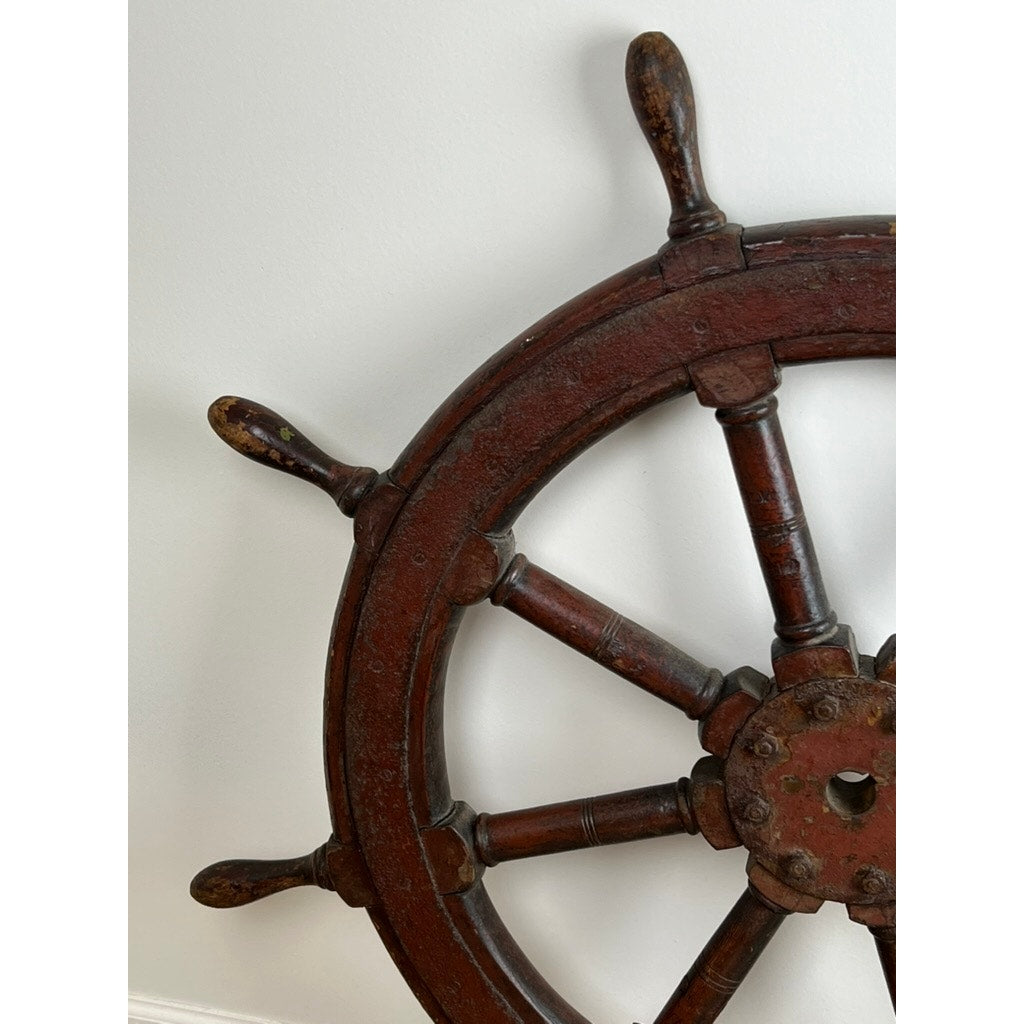 A close-up of the Antique Eight Spoke Mahogany and Iron Ship’s Wheel stamped “1864 US” shows rich patina and age, making it a striking piece of 19th-century maritime history displayed against a white wall.