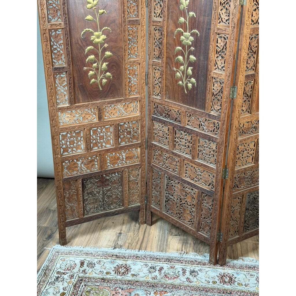 An Early 20th Century Anglo-Indian Carved Teak Four-Panel Screen with Applied Brass Floral Decoration from Pearl City Antiques stands on a wooden floor beside an area rug featuring beige and light blue patterns.