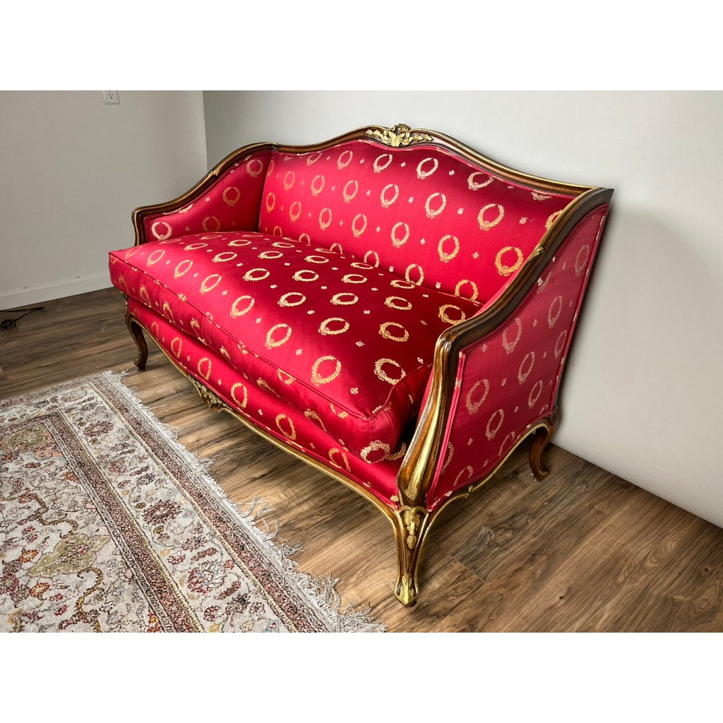 A Vintage French Regency Style Custom Upholstered Settee with red fabric and gold trim stands on a wooden floor beside a patterned area rug and against a white wall.
