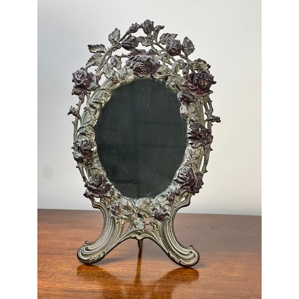 The Antique Art Nouveau Silvered Cast Brass Rose Vanity Dressing Mirror, dated 1909, features intricate rose and leaf designs on its decorative metal frame and stands on a wooden surface against a plain background.