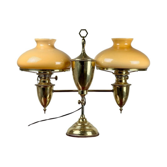 The Bradley and Hubbard Antique Brass Double-arm Student Lamp features carmel glass shades, an ornate central column, and an electric cord, shown against a white background.