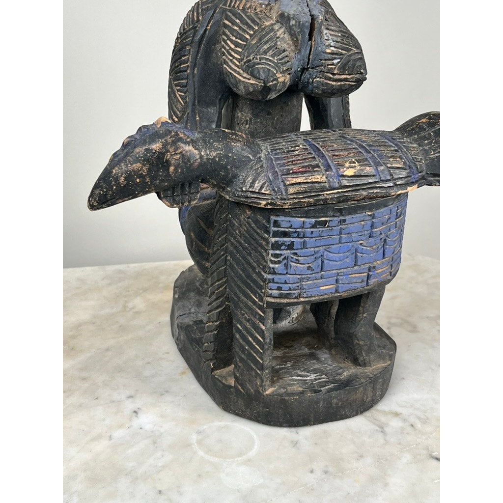 An Antique Igbo Ikenga Shrine Figure, probably from Nigeria, features a seated, bare-breasted figure with intricate patterns, holding a blue-painted animal-shaped object and displayed on a marble surface. Brand: Unknown.