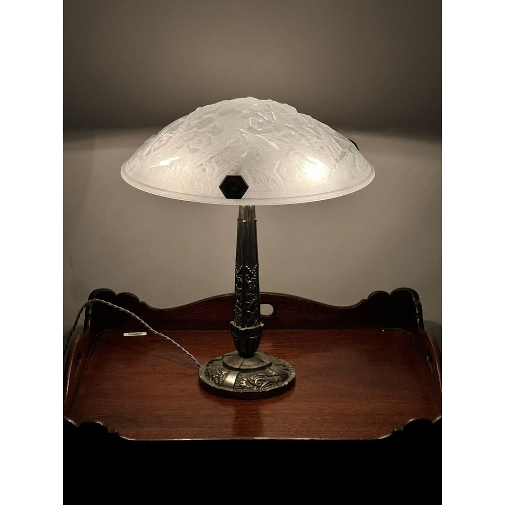 A Vintage French Art Deco frosted glass table lamp by Muller Frères Lunéville c. 1920, featuring geometric patterns, sits on a dark wooden tray table and casts a gentle glow over the area.