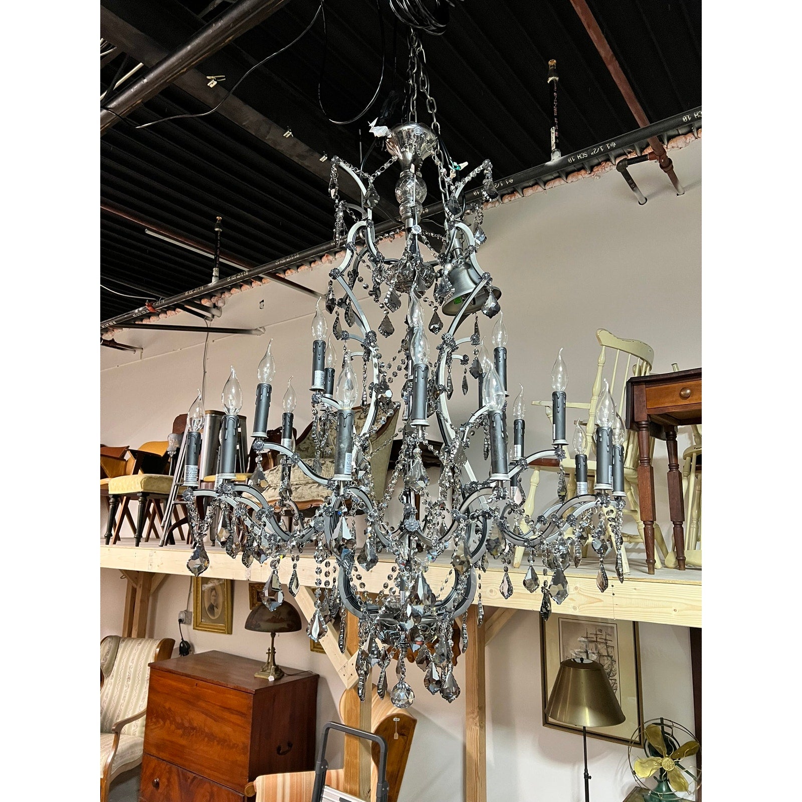 A Large Vintage Smoke Crystal 24 Light Chandelier hangs from the ceiling in a warehouse filled with assorted furniture and decor. Brand: None/Unknown.