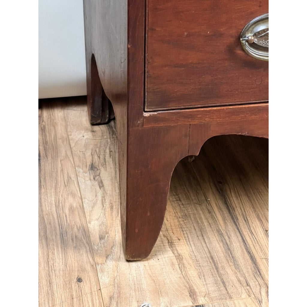 Corner of a Rare Labeled William Lloyd Springfield Massachusetts Federal Cherry Chest of Drawers, Circa 1800, showing its graceful curved leg and metal drawer pull as it stands on a wood floor. Evocative of early American craftsmanship, this piece represents the fine furniture created by cabinetmakers like William Lloyd in the Federal period. A portion of a white appliance is visible in the background.