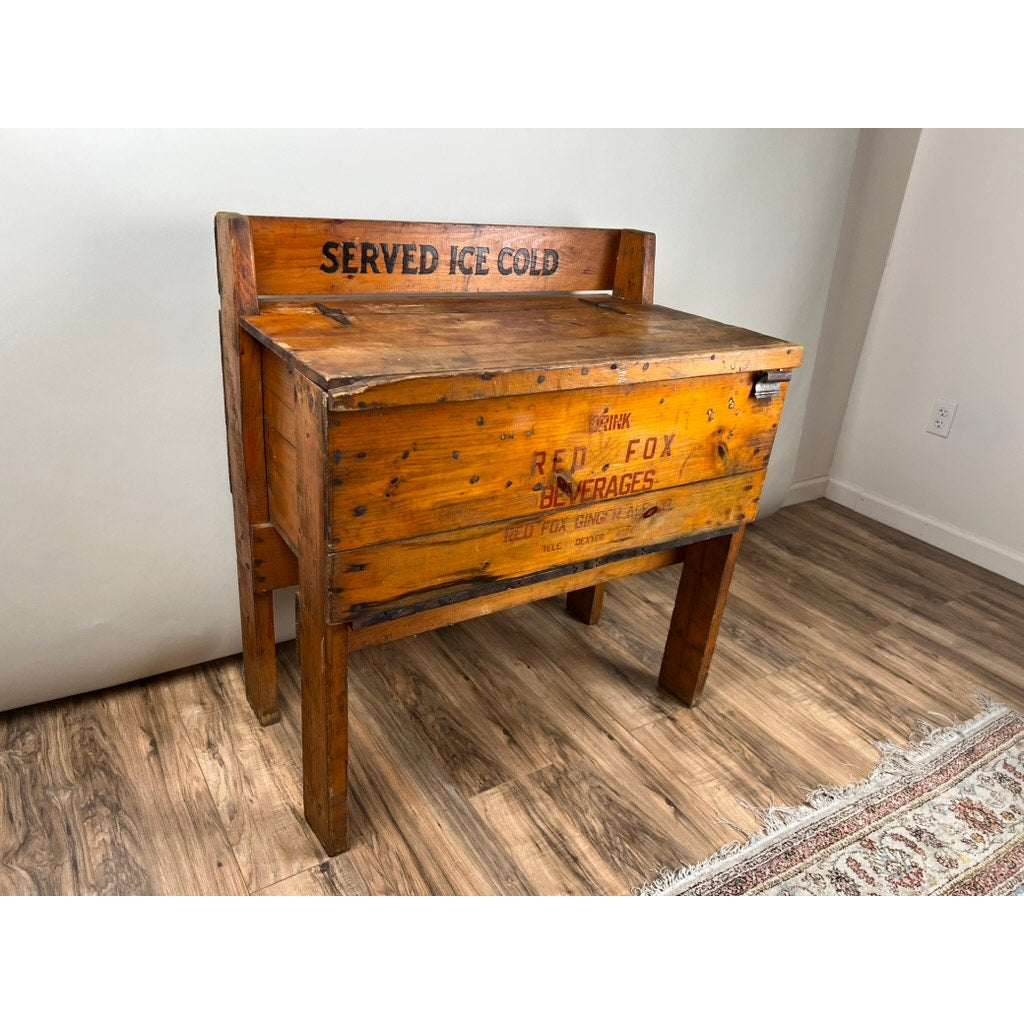 An Antique Tin Lined Pine Red Fox Ginger Ale of Rhode Island Cooler c. 1930, by Red Fox, stands on wooden legs with "SERVED ICE COLD" painted on front—perfect for accenting a hardwood floor and patterned rug.