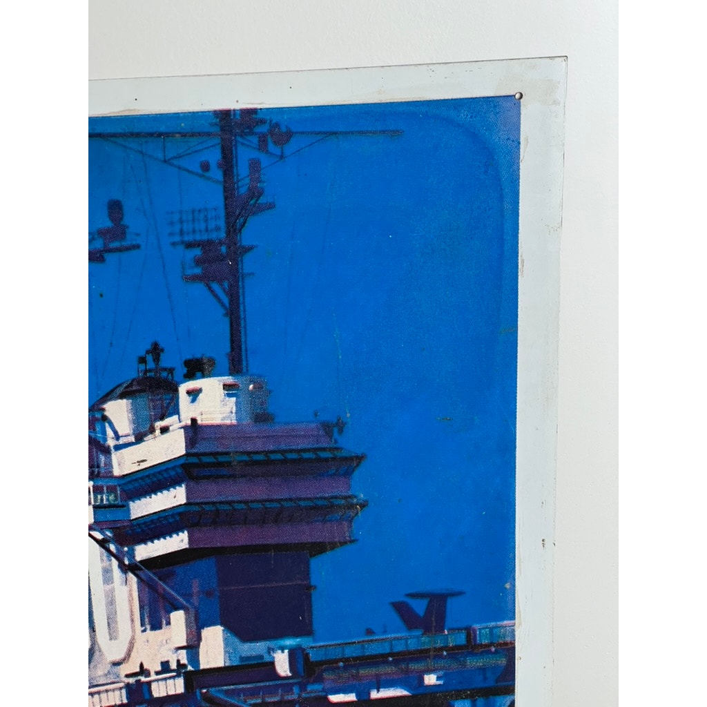 A close-up photo shows part of an original Vietnam Era double sided U.S. Navy recruiting tin sign by Lou Nolan, featuring the USS Saratoga. This collectible artwork is blue-tinted and pinned to a white wall.