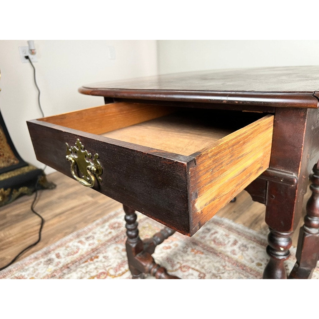 A close-up of an Antique English William & Mary Walnut One Drawer Gateleg Dining Table c. Early 18th Century shows its partially open drawer with a brass handle, revealing the empty interior and turned legs over a patterned rug.