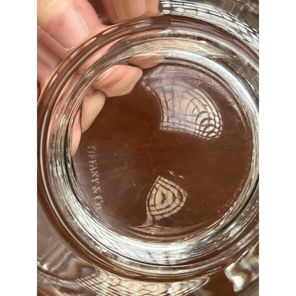 A close-up of the base of a Vintage Tiffany & Co. Mid-Century Modern Crystal Dolphin Pattern Footed Bowl shows the engraved TIFFANY & CO. logo, with reflections of a hand and a patterned window above.