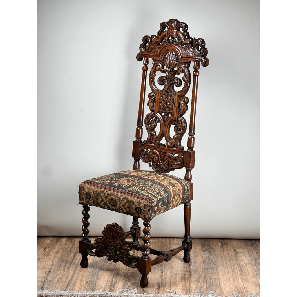 The Antique Carolean Revival Side Chair (late 19th c.), of unknown brand, features intricate carvings on its tall backrest and legs and a patterned tapestry-style upholstered seat. It is displayed on a wooden floor against a plain light background.