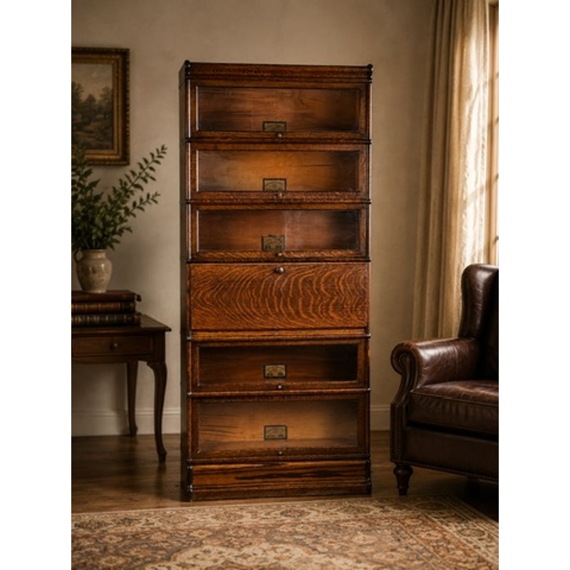 The Antique Globe-Wernicke Quarter Sawn Oak 6-Stack Barrister Bookcase Secretary Desk, crafted circa 1905–1915 by Globe Wernicke, features six sections each fitted with a glass-fronted door. The stately piece stands in a warmly lit room alongside a brown leather chair, a table stacked with books, a framed picture, and beige curtains.