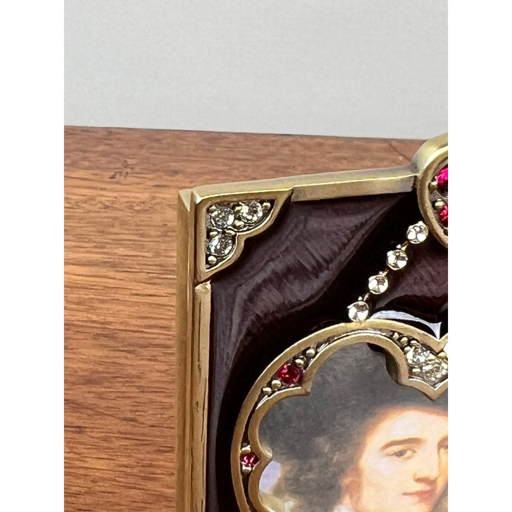 A close-up of the New in Box Jay Strongwater Bejeweled Enamel Double Heart Picture Frame shows its gold trim, sparkling jewels, and part of a painted portrait, set against a wooden background.