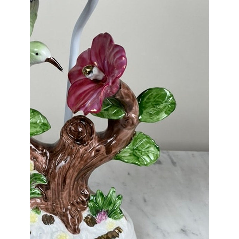A close-up of a Vintage Lena Liu Porcelain Hummingbird Accent Lamp, featuring a brown tree trunk, green leaves, and a large pink flower set on a white marble surface against a plain beige background.
