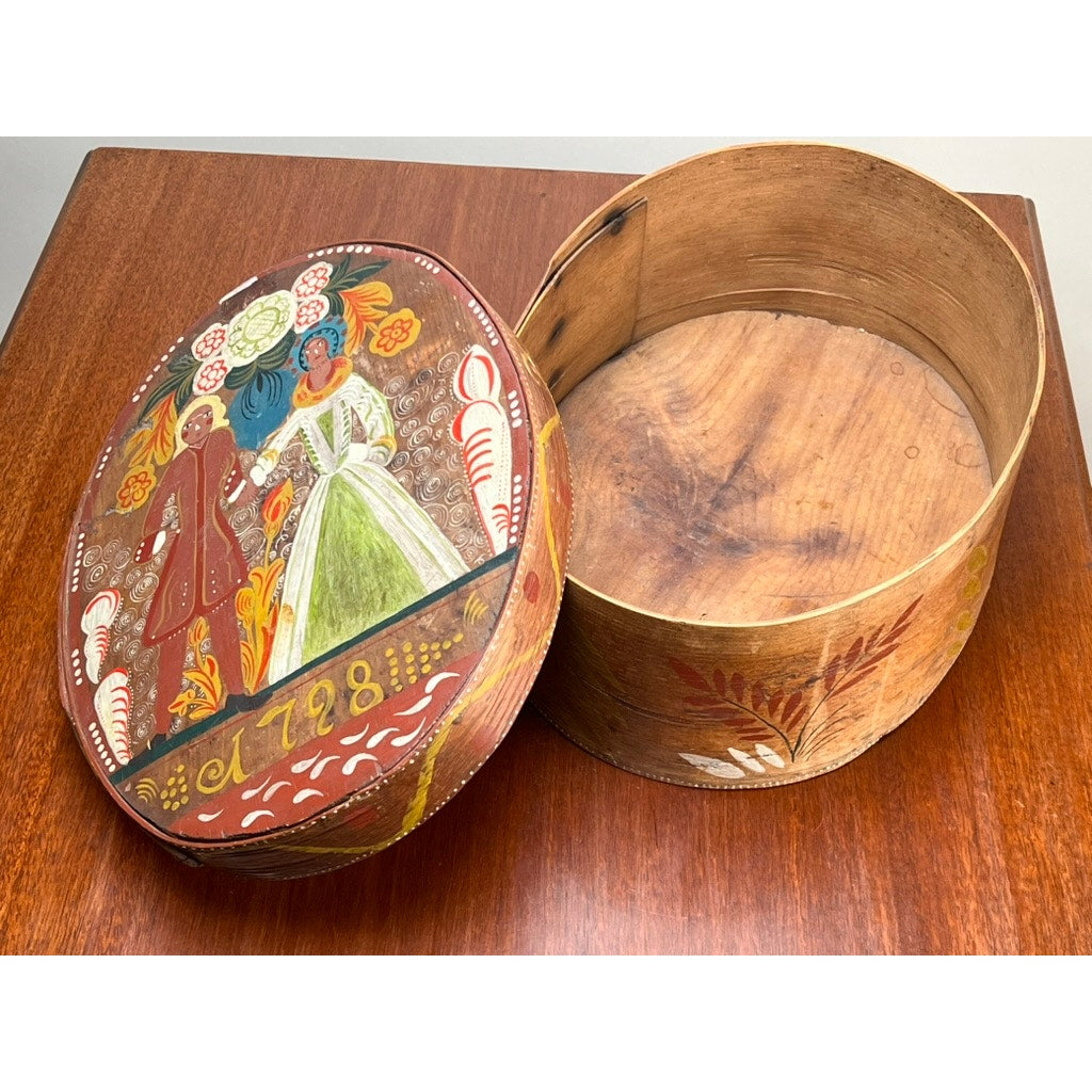 An Antique Swedish Polychrome Painted Marriage Box dated 1798 with bentwood construction and folk art motifs rests on a wooden surface, its lid showing a couple in traditional dress, floral designs, and the year 1798. Brand unknown.