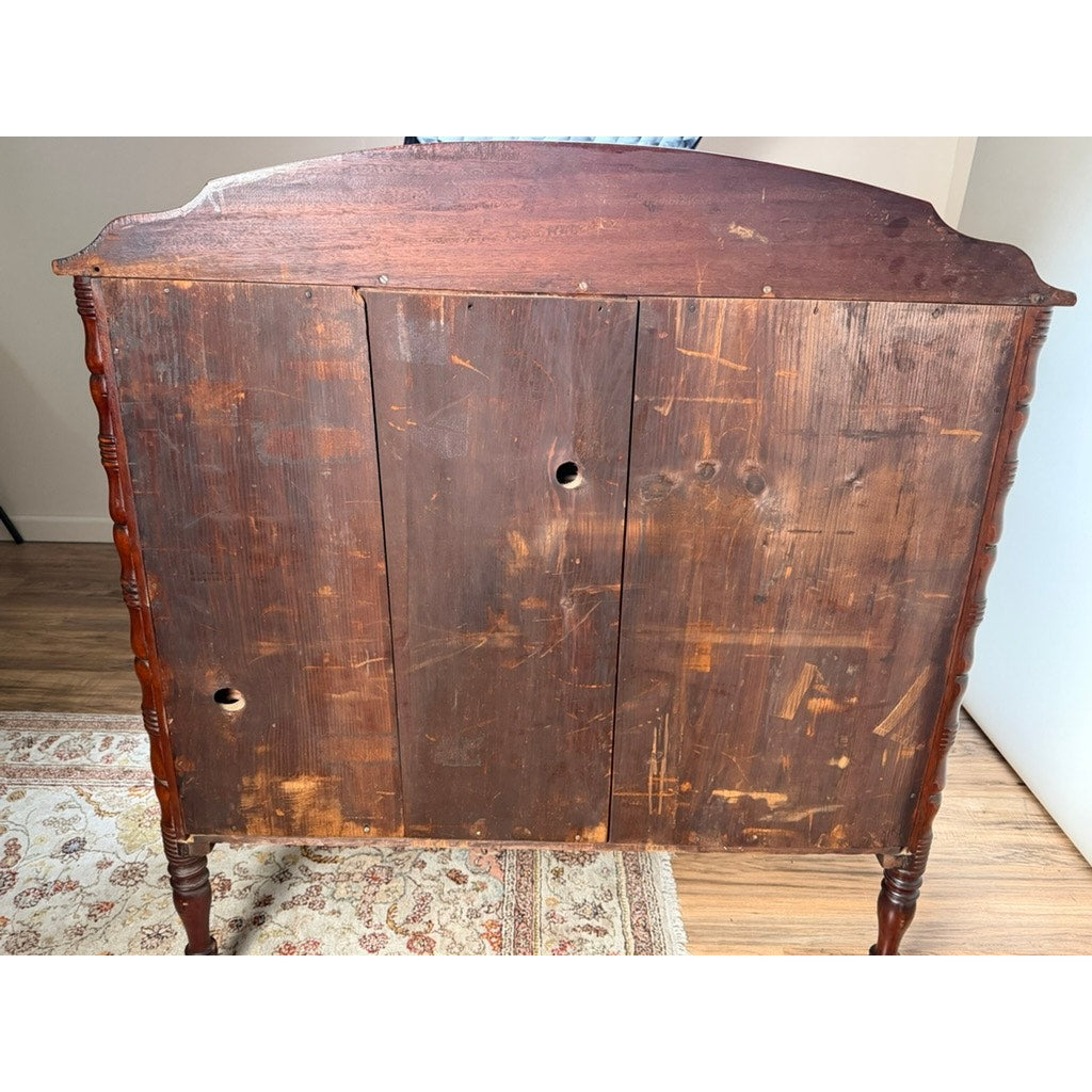 An Early 19th Century New England Federal Mahogany Bow-Front Chest of Drawers with Backsplash, dating from around 1820–1840, featuring three paneled sections, visible scratches, two holes, spindle legs, and a curved top, stands on a hardwood floor next to a patterned rug.