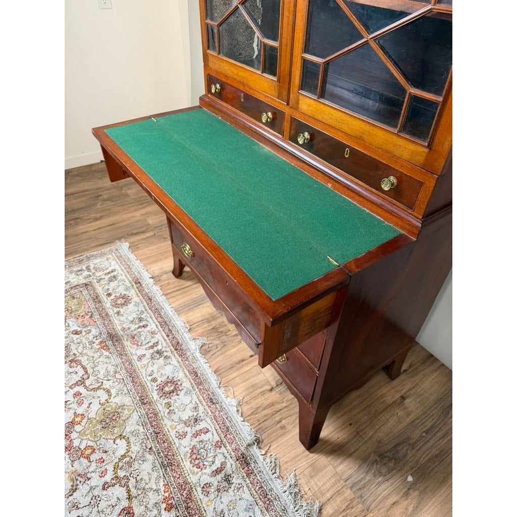 A Federal Flame Mahogany Secretary Bookcase with Eagle Finial, dating to circa 1810–1825, featuring a green felt writing surface when open, glass-front cabinets above, and drawers below—all set atop a hardwood floor with a glimpse of patterned rug—an exemplary piece of early 19th-century American cabinetmaking.