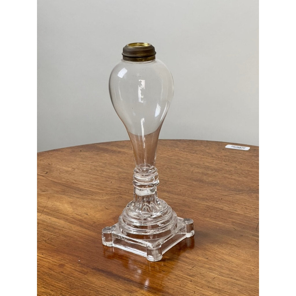 An Attributed Boston & Sandwich Pressed Glass Boudoir Fluid Lamp, circa 1850, featuring clear glass with a rounded top and brass fitting atop an ornate square base, stands on a wooden table against a plain light background—an elegant example of early American lighting design.