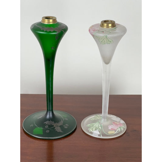 A pair of antique Art Nouveau fluted oil lamps from Pearl City Antiques, circa 1920, are displayed on a wooden surface. The left lamp features green glass with etched floral designs, evoking vintage charm, while the right lamp is crafted from frosted glass adorned with hand-painted pink and green flowers. Both lamps are topped with elegant brass candle holders, enhancing their timeless appeal as home decor accents.