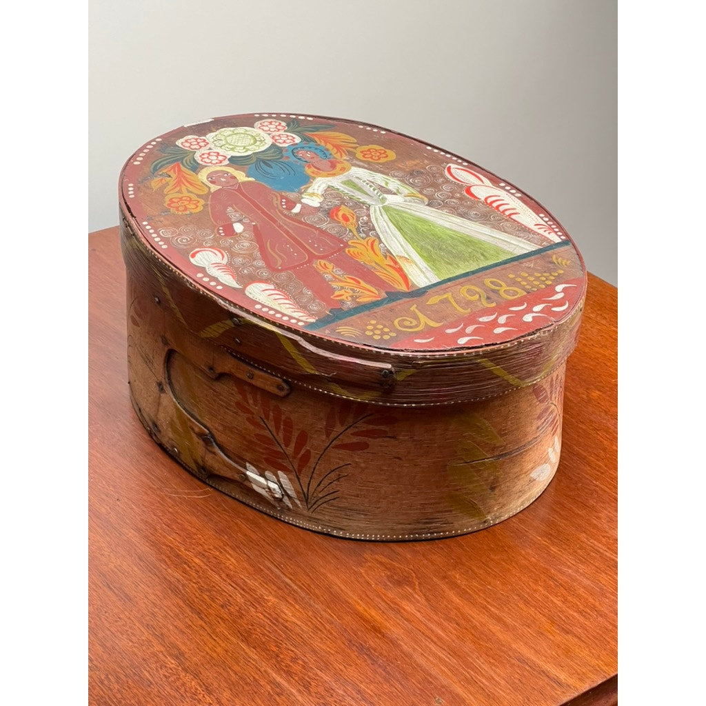 The Antique Swedish Polychrome Painted Marriage Box dated 1798 is an oval bentwood box featuring folk art with floral patterns and figures in traditional dress, displayed on a wooden table against a plain wall. Brand: None/Unknown.