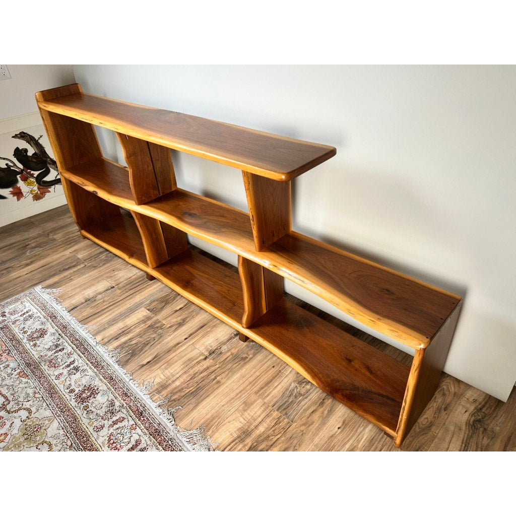 A Vintage Mid-Century Modern Style Live Edge Walnut Bookshelf with multiple open shelves and a natural wood grain stands against a white wall on a wood floor, beside a patterned rug. Brand: None and/or Unknown.