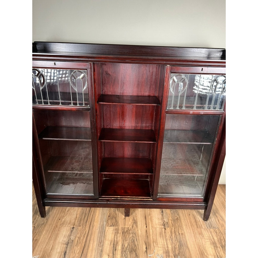 The Antique Arts & Crafts Leaded Glass Mahogany Bookcase c. 1920 features glass doors with decorative leaded glass patterns at the top, open center shelves, and stands on short legs, perfect for display on a wooden floor.