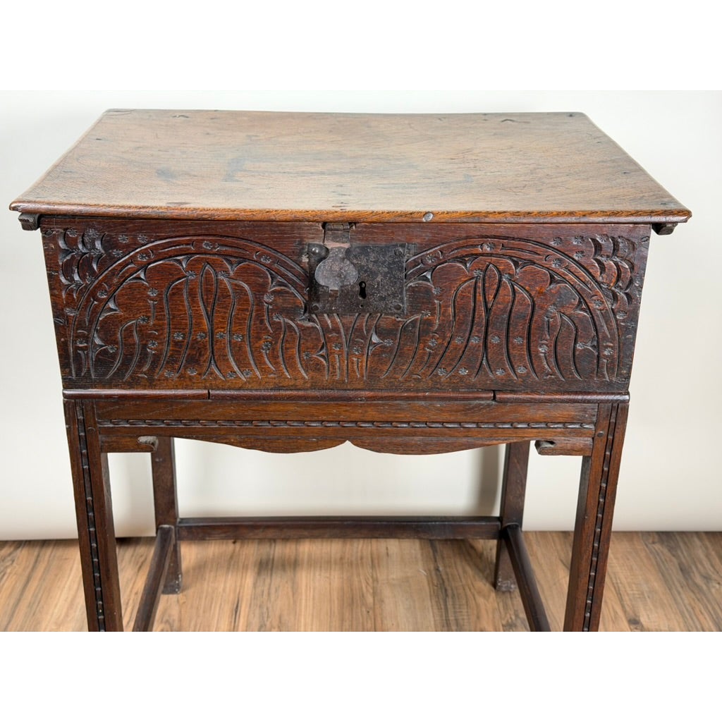 An English Jacobean Carved Oak Bible Box on Later Stand from Pearl City Antiques, featuring intricate carved front panels and a metal latch, resting on a four-legged stand.