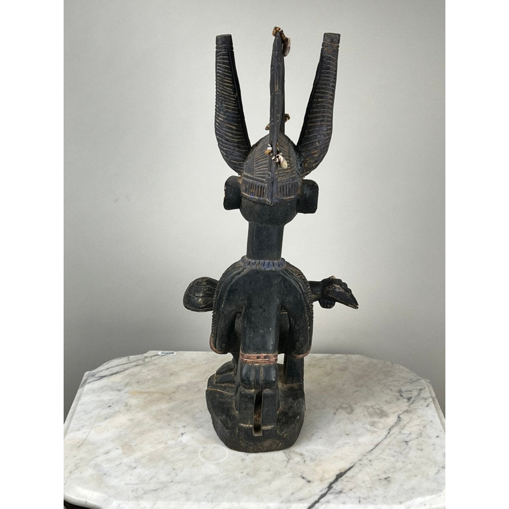 An Antique Igbo Ikenga Shrine Figure, probably from Nigeria, features tall curved horns and decorative carvings. Viewed from the back, the wooden sculpture sits on a marble surface against a plain light gray background. Brand: Unknown.
