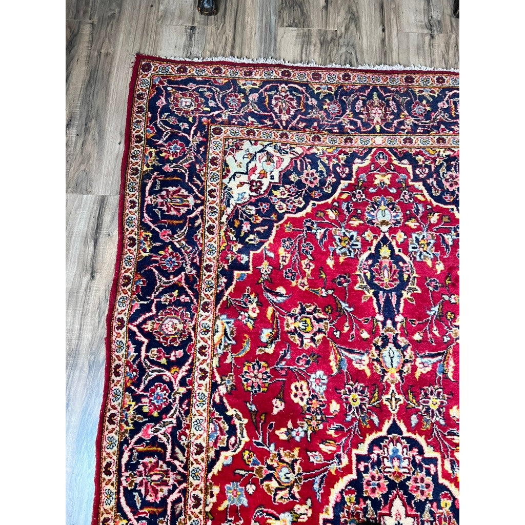Corner of a Vintage Hand-Knotted Persian Kashan Wool Gallery Rug (c. 1950–1970, 4’5 x 8’8) with intricate red, navy, cream, and yellow floral patterns and detailed border, displayed on a wooden floor. Brand: None/Unknown.