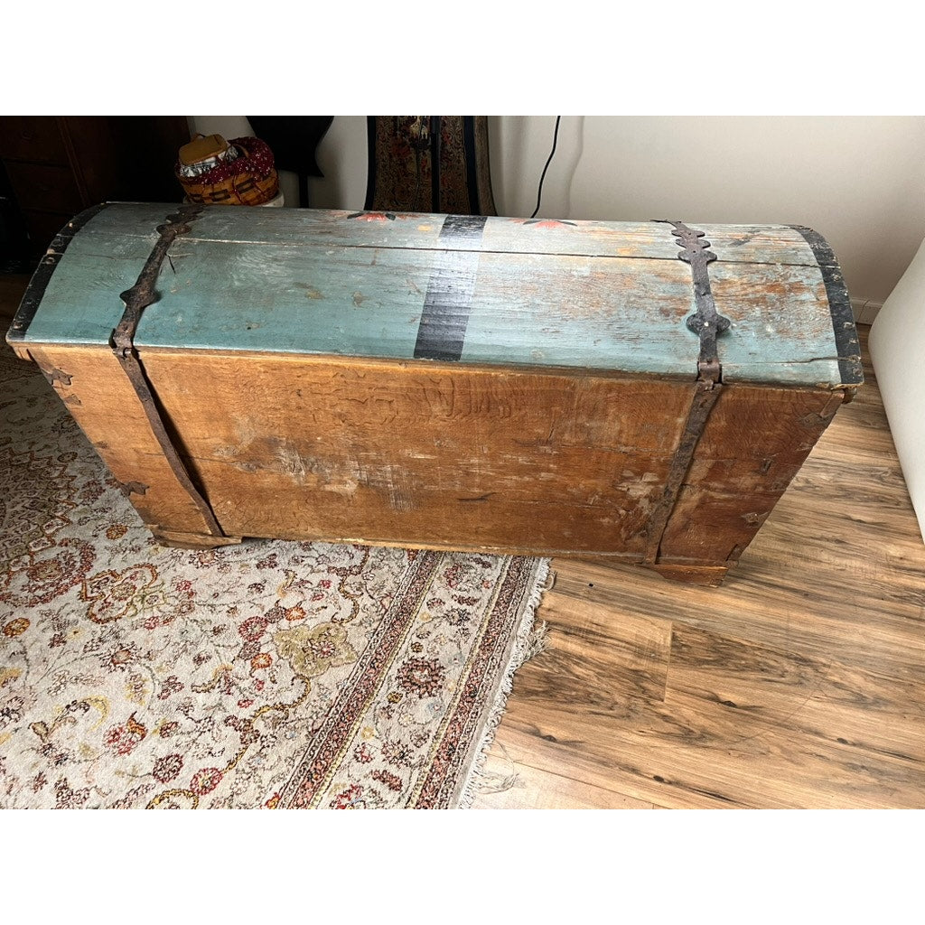 The Antique Swedish Oak Kista Dowry Chest c. 1841, with its curved blue top and rustic metal bands, sits on a wooden floor by a patterned rug, embodying the timeless charm of Swedish folk art.