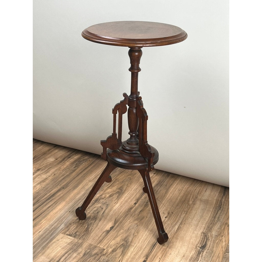 An Antique Victorian Walnut Candle Stand from the late 19th century, featuring a round top, ornate central column, and three curved legs, is displayed on a wooden floor against a plain light background. Brand: None/Unknown.