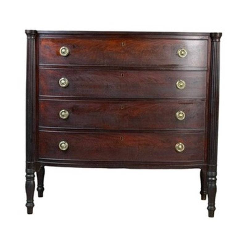 This Early 19th Century New England Federal Mahogany Bow-Front Chest of Drawers (c. 1810–1825) features four gracefully curved drawers, each fitted with two round brass handles. Slender turned legs and a rich dark brown finish highlight the timeless elegance characteristic of Federal-era furniture.