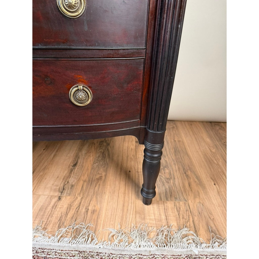 Close-up of the lower corner of an early 19th Century New England Federal mahogany bow-front chest of drawers (c. 1810–1825), featuring brass drawer pulls, standing on a wooden floor beside the fringed edge of a rug.