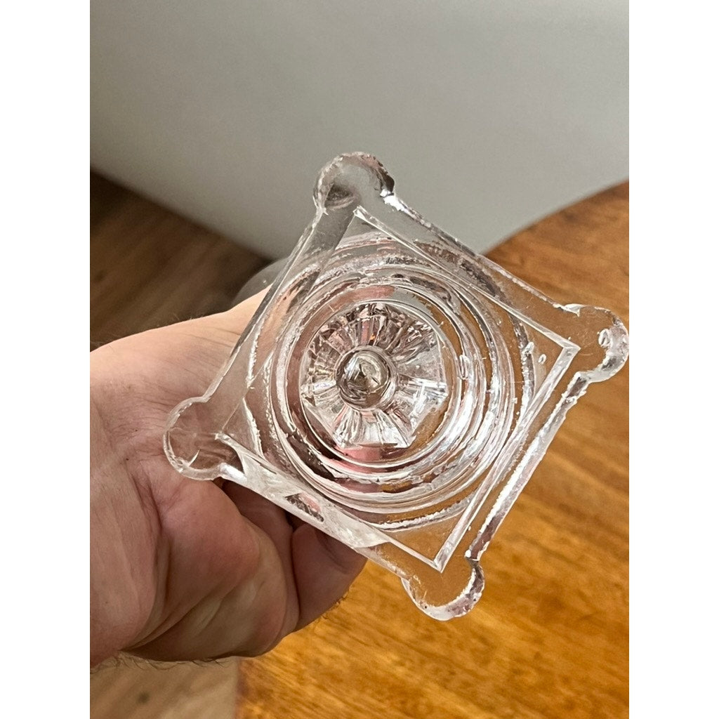 A hand holds an Attributed Boston & Sandwich Pressed Glass Boudoir Fluid Lamp, Circa 1850, featuring a clear glass construction with a square, decorative base and a circular, patterned center reminiscent of early American lighting. A wooden surface and gray background are visible.