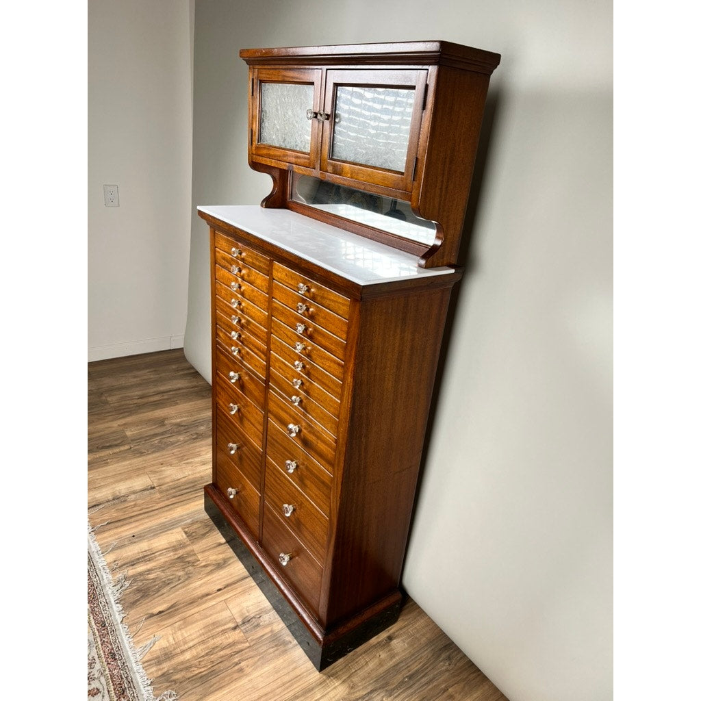 An Antique Mahogany Multi-drawer Dental Cabinet by The American Cabinet Co. (early 20th century) features a white countertop, numerous small drawers with metal handles, frosted glass upper doors, and a mirrored backsplash against a light wall.