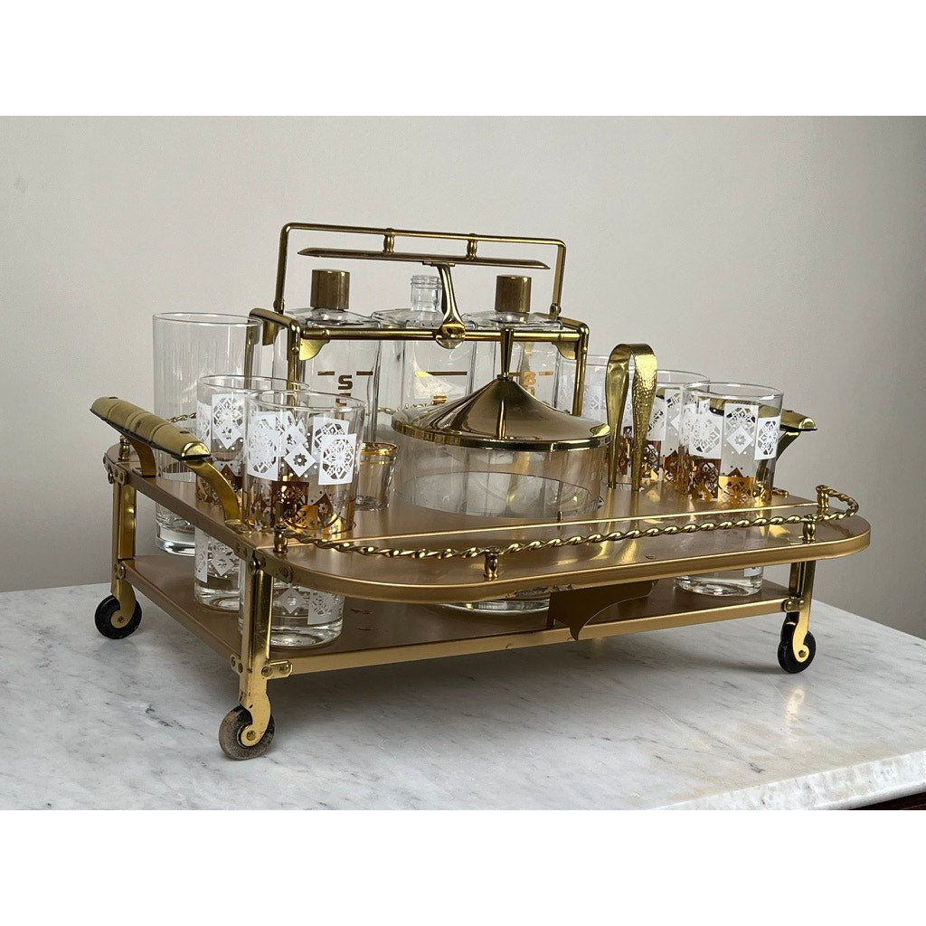 A Vintage Mid Century Modern Bar Caddy c. 1960s holds glassware, including a decanter, gold-detailed glasses, and a pitcher. It sits on a white marble surface against a plain light-colored wall. Brand: None/Unknown.
