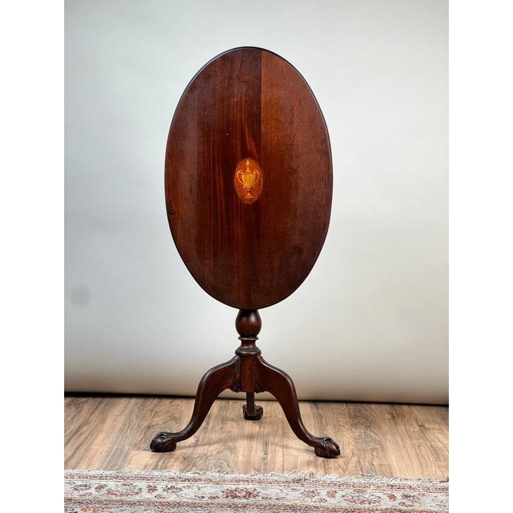 The Antique George III Tilt-top Tea Table by Pearl City Antiques, crafted from dark mahogany with an oval top and accented by a central satinwood inlay, features a tripod pedestal base ending in intricately carved claw feet. It is displayed on a wooden floor with part of a rug visible beneath.