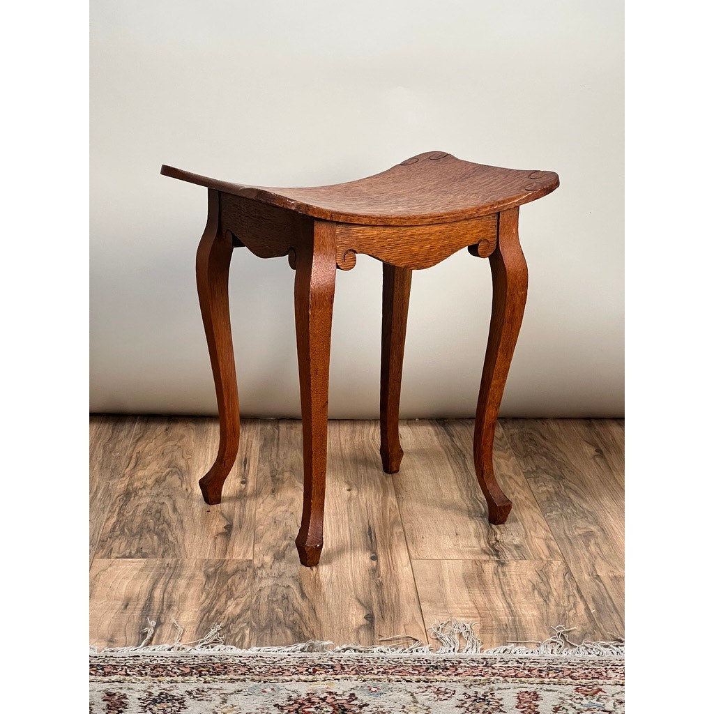 Antique Arts and Crafts Saddle Shaped Footstool c. 1900 features a curved saddle seat and four elegantly curved legs, displayed on a wooden floor with a patterned rug partially visible. Brand: None/Unknown.