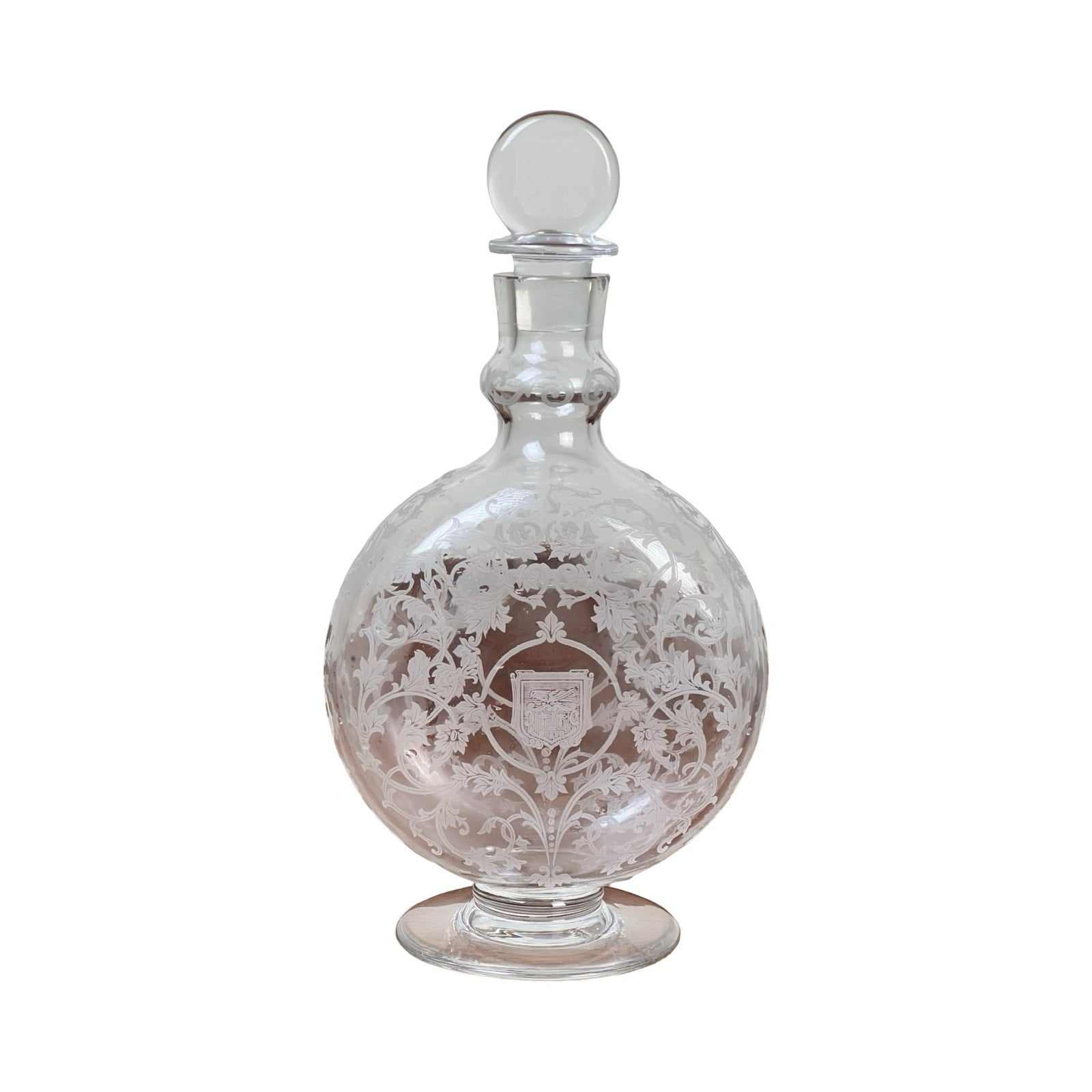 A vintage Baccarat "Michelangelo" crystal decanter for Martell Cognac c. 1930, featuring ornate floral and leaf engraving, a stopper, a small shield design on the front, and a round clear body on a flat circular base.
