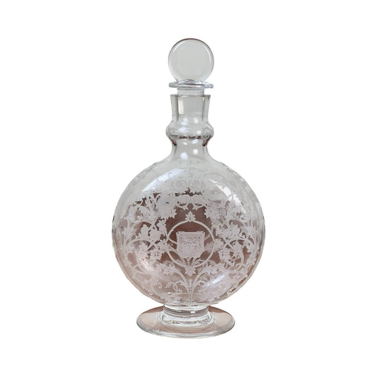 A vintage Baccarat "Michelangelo" crystal decanter for Martell Cognac c. 1930, featuring ornate floral and leaf engraving, a stopper, a small shield design on the front, and a round clear body on a flat circular base.