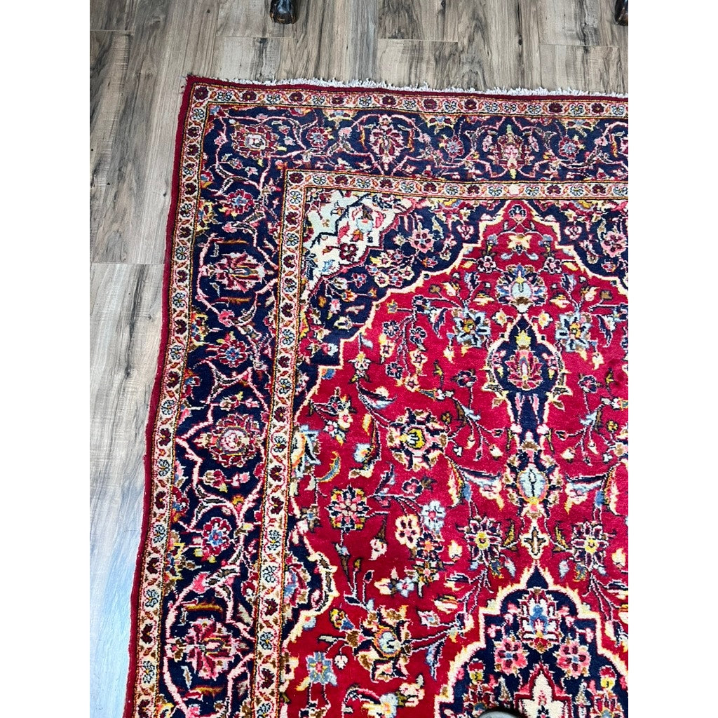 A corner of a Vintage Hand-Knotted Persian Kashan Wool Gallery Rug (c. 1950–1970, 4’5 x 8’8) with intricate red, navy blue, yellow, and white floral patterns rests on a wooden floor; its edge is slightly uneven.