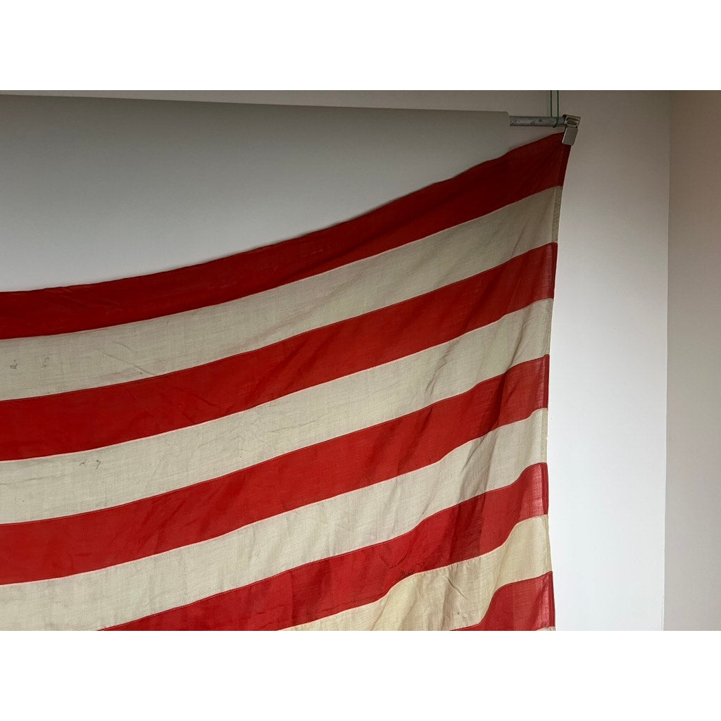 A partial view of an antique 45-star American flag from the Spanish–American War era (9’9 x 6), featuring red and white stripes, hangs from a rod against a white wall. Brand: None/Unknown.