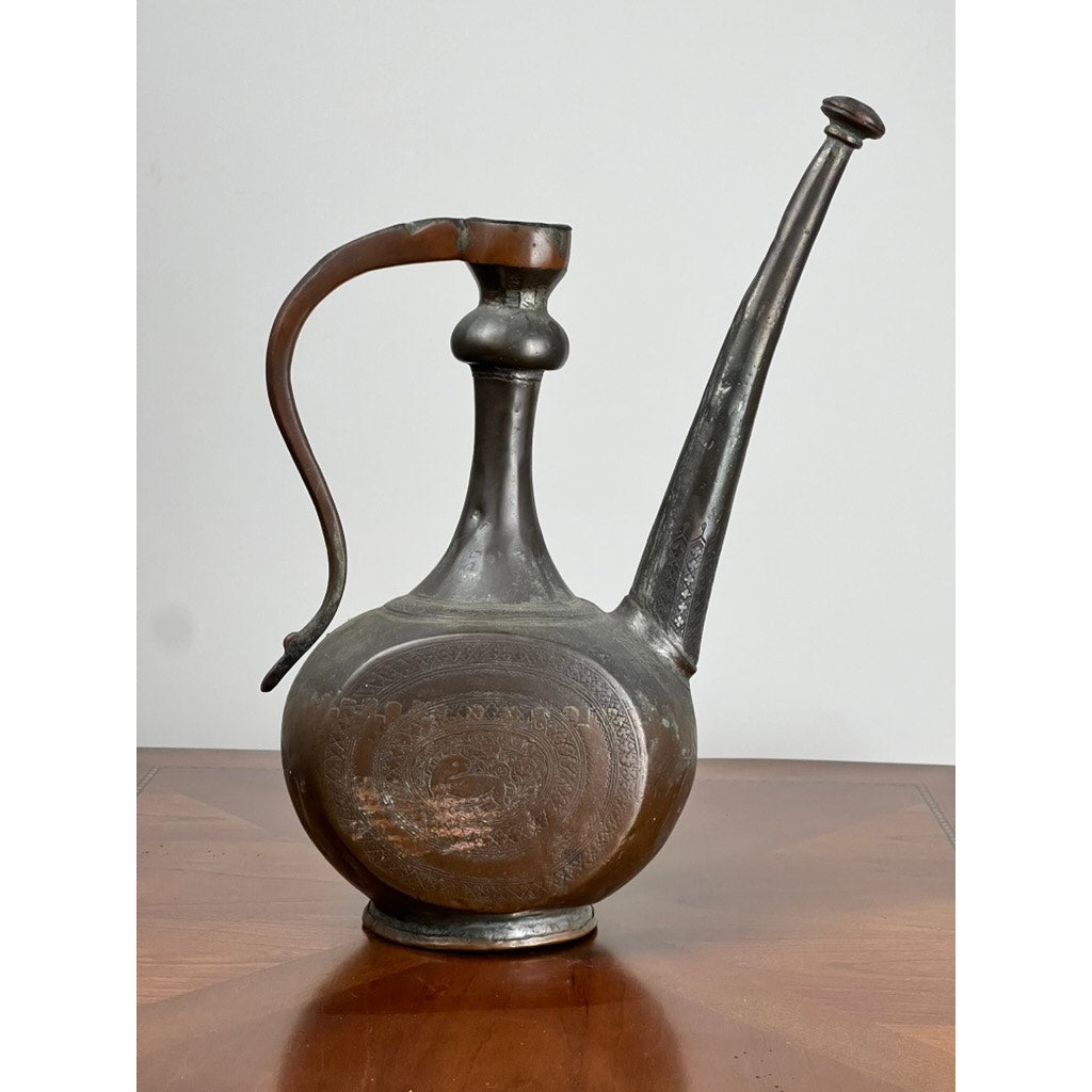 An Antique Qajar Persian Copper Engraved Ewer (19th Century) with a long spout, curved handle, and decorative patterns highlights classic Persian metalwork, displayed on a wooden surface against a plain light background.