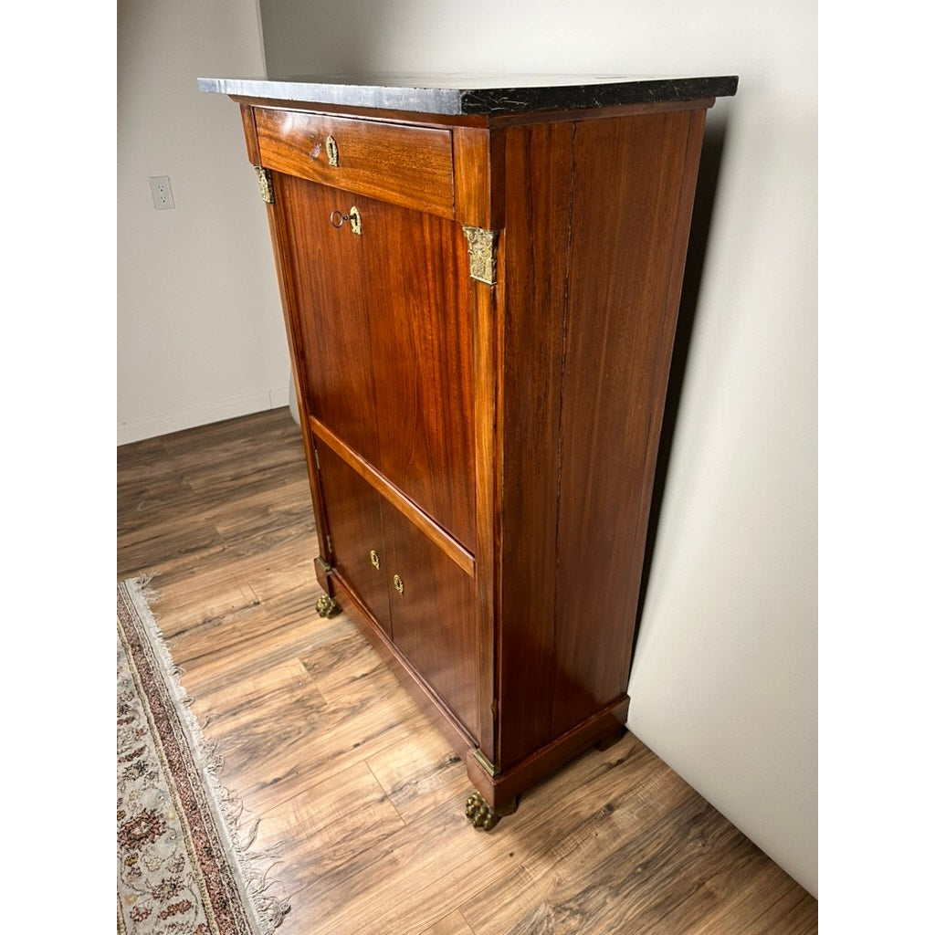 An Antique French Empire Secrétaire Abattant with Ormolu Mounts, c. mid-19th century, featuring mahogany veneer and a dark marble top, stands on a wood floor near a patterned rug and light-colored wall.